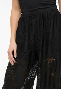 Czarne spodnie z koronki z kwiatowym wzorem, wyposażone w elastyczną talię i luźny, zwiewny krój, podkreślające przezroczystość w niektórych miejscach.