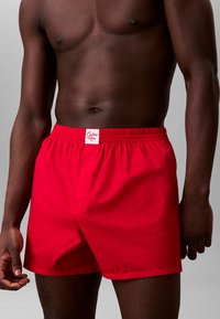 Costume da bagno rosso con taglio di lunghezza media, cintura elastica e una piccola etichetta con il marchio sul davanti. Tessuto morbido, design minimale.