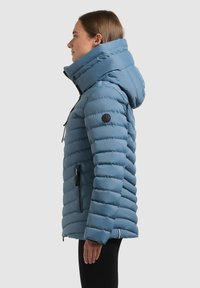 Giacca piumino azzurro chiaro con design a righe orizzontali, dotata di cappuccio, tasche con zip e un logo sulla manica.