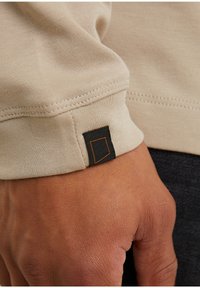Beige Langarmshirt-Manschette aus strukturiertem Stoff; mit einem schwarzen Etikett mit orangefarbenem Rand. Nahaufnahme, die die Nähdetails betont.