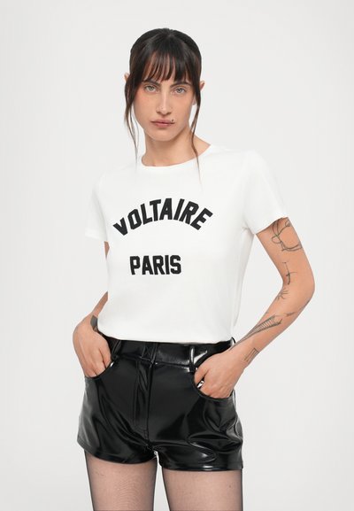 Weißes T-Shirt mit kurzen Ärmeln und schwarzem "VOLTAIRE PARIS" Text. Kombiniert mit hoch taillierten, glanz schwarzen Shorts. Schlichter Hintergrund.