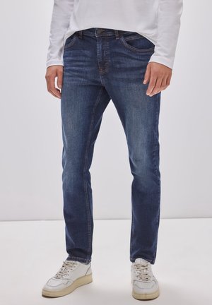 Homme portant un jean slim bleu foncé, une chemise blanche à manches longues et des baskets blanches, debout sur un fond clair uni.