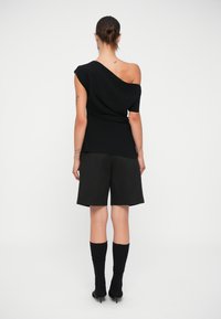 Donna in piedi di spalle, con indosso un top nero asimmetrico, pantaloni corti neri fino al ginocchio, stivali alti neri, e tatuaggi su braccia e collo.