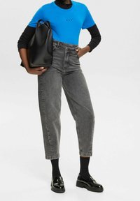 Blå T-shirt med sorte detaljer, højtaljede grå jeans og sorte loafers. Sort taske med en glat tekstur, stående på en ensfarvet baggrund.