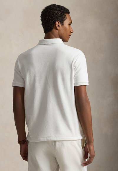 Mężczyzna z krótkimi kręconymi włosami, ubrany w gładką białą koszulkę polo z krótkim rękawem i białe spodnie, stojący tyłem na neutralnym tle.