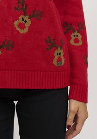 Pull tricoté rouge avec des motifs de rennes en marron et beige. Poignets et ourlet côtelés. Accent en métal près du bas.