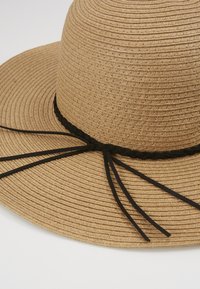 Breitkrempiger Strohhut in natürlichem Beige mit strukturierter Oberfläche. Mit einem schwarzen geflochtenen Band und zwei schwarzen hängenden Bändern.