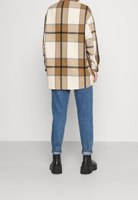 Camicia quadrata oversize nei colori beige, marrone e nero, realizzata in tessuto morbido, abbinata a jeans a gamba affusolata blu chiaro e stivali neri alla caviglia.
