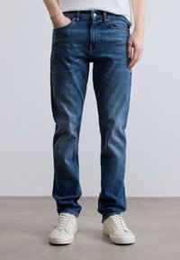 Plave traperice klasičnog kroja, s pet džepova i diskretnim izblijedjelim dijelovima. Položene ravno uz bijele tenisice, prikazujući ležerni dizajn.