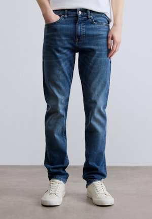 Modré džínsové nohavice klasického strihu s piatimi vreckami a jemným vyblednutím. Položené na rovnej ploche spolu s bielymi teniskami, ktoré zobrazujú ležérny dizajn.