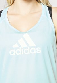 Jasnoniebieski sportowy top na ramiączkach z dekoltem w serek, wykonany z lekkiego materiału, z białym logo Adidas na przodzie i czarnymi akcentami na ramiączkach.