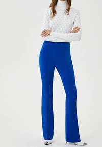 Blaue Schlaghosen aus einem dehnbaren Stoff, mit einem breiten Bund und glatter Oberfläche. Kombiniert mit einem weißen, strukturierten Rollkragenoberteil.