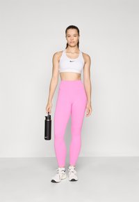 White sports bra with black logo, high-waisted pink leggings, black water bottle, and white sneakers with gray accents, standing against a light background.