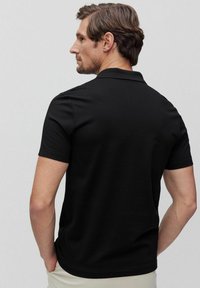 Svart kortärmad polo med krage, slät textur och en figurnära passform, sedd bakifrån. Minimalistisk design utan synliga accenter.
