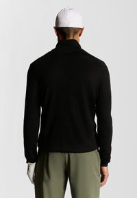 Pullover in pile nero con colletto alto, vista posteriore. Caratterizzato da maniche lunghe e una vestibilità rilassata. Indossato con pantaloni verde oliva e un cappellino bianco.
