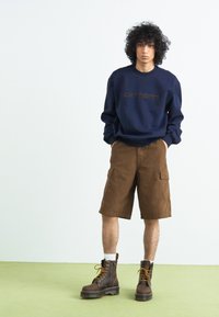 Navy sweatshirt with embossed brand logo, brown cargo shorts with pockets, and brown lace-up boots, standing on light green flooring.