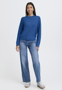 Pull en tricot bleu avec un design côtelé, col rond et manches longues, associé à un jean bleu clair ample et des baskets grises.