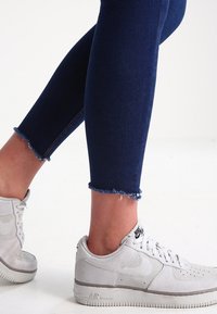 Vaqueros ajustados azul oscuro con bajos deshilachados, combinados con zapatillas Nike blancas texturizadas con un acento gris y diseño perforado.