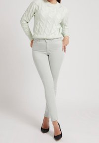 Kvinna i ljusgrå skinny jeans, vit kabelstickad tröja och svarta spetsiga högklackade skor, stående med korslagda ben.