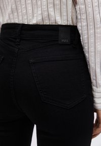 Oseba, ki nosi črne kavbojke z zadnjim žepom in črno usnjeno etiketo z napisom "BOSS", združene s svetlo črtasto srajco.