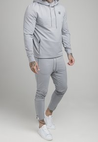 Hombre con sudadera gris claro y joggers a juego, con zapatillas blancas, tatuajes visibles en las manos y tobillos, de pie sobre un fondo gris.