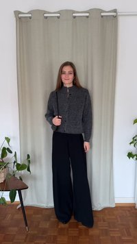 Grå strukturert cardigan med høy krage, svarte vide bukser, og en liten svart skulderveske, plassert foran et lyst gardinbakgrunn.