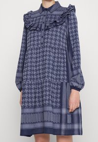 Granatowa sukienka w wzór houndstooth i geometryczny, z falbaniastego kołnierza, guziki z przodu oraz długimi bufkami z elastycznymi mankietami.