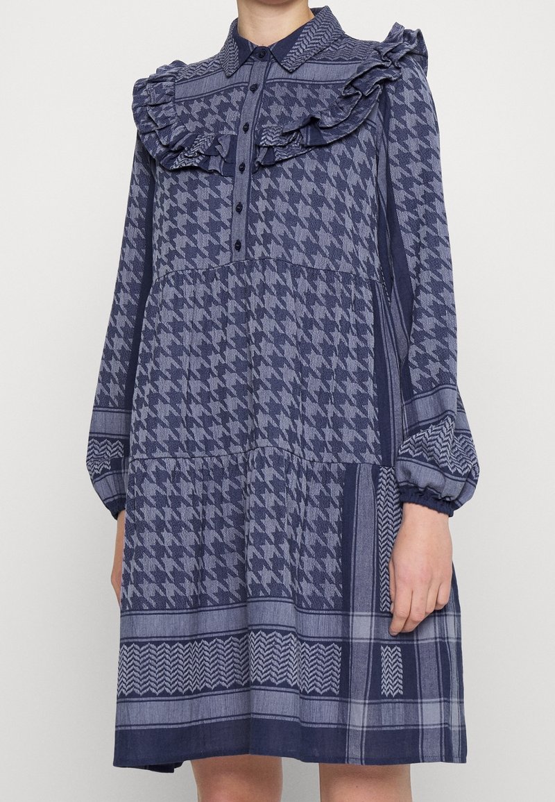 Granatowa sukienka w wzór houndstooth i geometryczny, z falbaniastego kołnierza, guziki z przodu oraz długimi bufkami z elastycznymi mankietami.