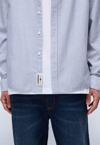 Chemise rayée bleue et blanche, design à boutons, en coton, avec une étiquette sur le côté et des manches longues. Portée par-dessus un t-shirt blanc et un jean foncé.
