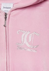 Pink velour zip-up jacket featuring silver glittery logo and text. The zipper pull is a looped design, and the fabric has a soft texture.