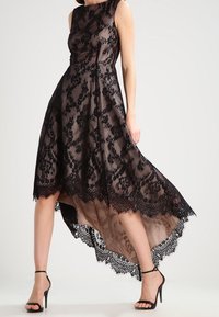 Svart spetsklänning med genomskinligt överlager, hög-låg fåll, ärmlös design och intrikata blomsterdetaljer längs kanterna. Svarta remklackar.