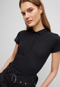 Schwarzes Poloshirt mit kurzen Ärmeln, glatter Textur, Knopfleiste und dezentem BOSS-Logo. Kombiniert mit hochtaillierten schwarzen Hosen.