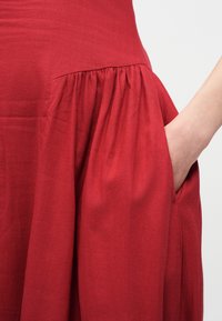 Χέρι που ξεκουράζεται μέσα στην τσέπη ενός κόκκινου φορέματος με ρίγες, πάνω σε απλό φωτεινό φόντο.