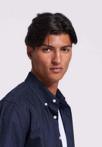 Man met donker haar in een marineblauw overhemd met knopen over een wit T-shirt, kijkt iets opzij tegen een effen lichte achtergrond.