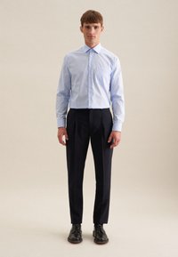 Camicia azzurra chiara con taschino, abbinata a pantaloni neri plissettati e scarpe nere stringate, su sfondo neutro.