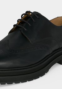Chaussure brogue en cuir noir avec perforations décoratives, bout rond, lacets noirs et semelle épaisse en caoutchouc avec motif de la semelle.