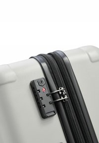 Valise rigide grise avec une fermeture éclair noire, un verrou à combinaison et une surface texturée. Conception durable avec une forme épurée.