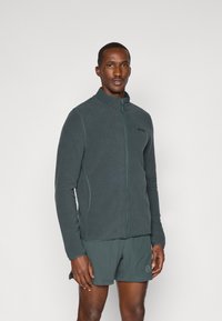 Ciemnozielona polarowa kurtka z wysokim kołnierzem, pełnym zamkiem błyskawicznym i kieszeniami bocznymi, noszona z dopasowanymi szortami. Posiada logo na klatce piersiowej.