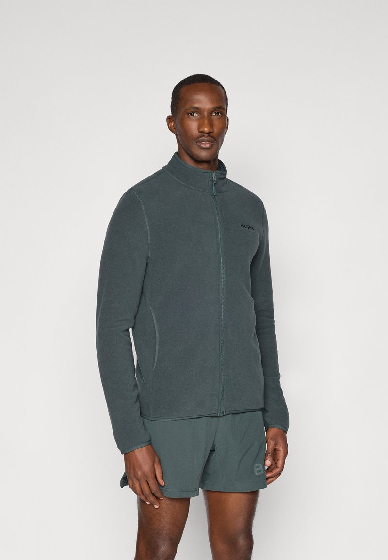 Ciemnozielona polarowa kurtka z wysokim kołnierzem, pełnym zamkiem błyskawicznym i kieszeniami bocznymi, noszona z dopasowanymi szortami. Posiada logo na klatce piersiowej.