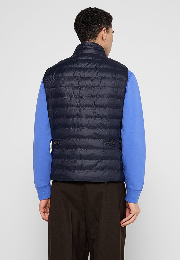 Granatowa kamizelka puchowa z poziomym pikowaniem, wysokim kołnierzem i dwoma guzikami przy talii, noszona na niebieskiej koszulce z długim rękawem.
