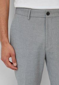 Grijze op maat gemaakte broek met een gestructureerde stof, voorzien van een knoopsluiting, ceintuurlussen en een vlakke voorkant. Hand rustend op het kledingstuk.
