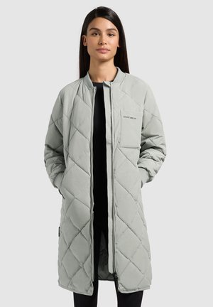 Moteris su ilgais tamsiais plaukais, dėvinti šviesiai pilką siūlytą paltą, siekiantį kelius, rankomis kišenėse, po juodu drabužiu.