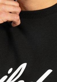 Czarny bawełniany t-shirt z okrągłym dekoltem, na froncie z białym napisem graficznym. Widać rękę trzymającą dekolt.