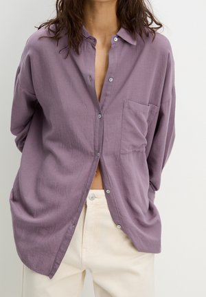 Femme portant une chemise ample violette à boutons avec poche avant et pantalon de couleur crème, mains derrière le dos, devant un fond uni.
