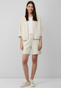 Hellbeige Zip-Jacke mit kurzen Ärmeln, kombiniert mit passenden Shorts. Beides aus weichem Stoff. Das Modell trägt beige Sneaker.