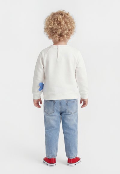Enfant aux cheveux blonds bouclés, debout de dos, portant une chemise blanche à manches longues, un jean bleu clair et des baskets rouges sur fond blanc.