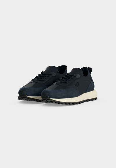 Paire de baskets de sport noires et bleu foncé à lacets, avec semelles intermédiaires blanches et semelles extérieures noires texturées sur fond uni.