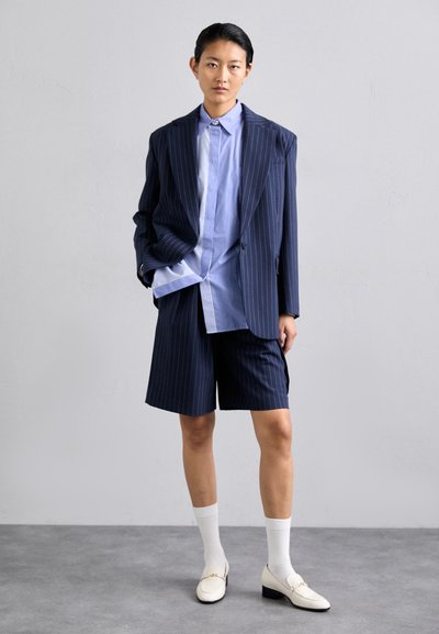Blazer en velours côtelé bleu marine sur une chemise rayée bleu clair et un short bleu marine, associé à des chaussettes blanches et des mocassins crème. Textures lisses tout au long.