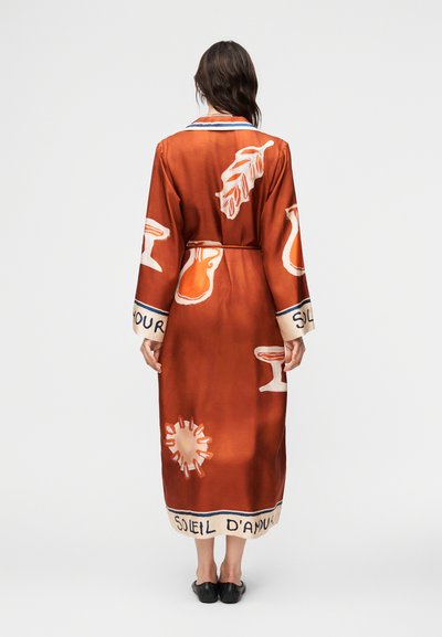 Γυναίκα που φοράει μακρύ φόρεμα σε σκουρόχρωμο πορτοκαλί με αφηρημένα λευκά και μπεζ σχέδια, όρθια και ξυπόλητη, γυρισμένη με την πλάτη προς το κοινό σε απλό φόντο.