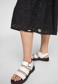 Sandales en cuir blanc avec plusieurs brides et un détail de boucle, associées à une robe noire texturée avec un motif floral.